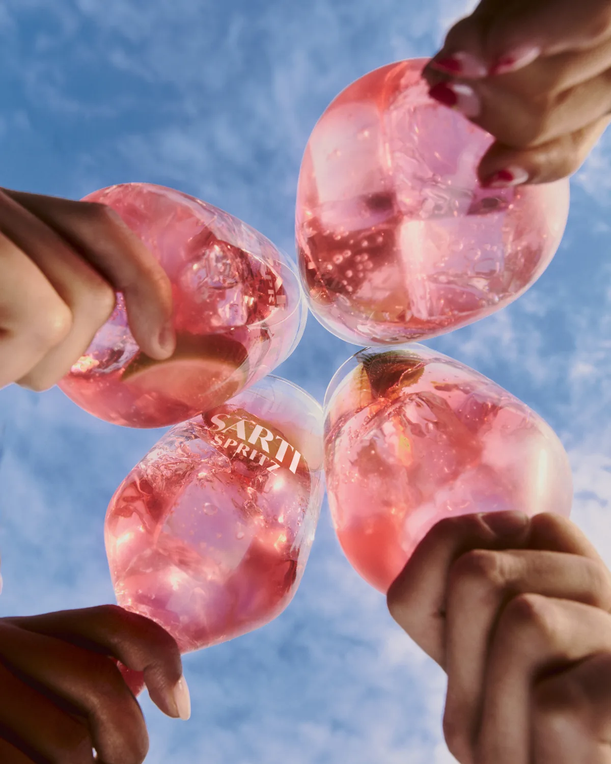 Quattro calici di Sarti Spritz che si toccano in un brindisi, visti dal basso contro il cielo