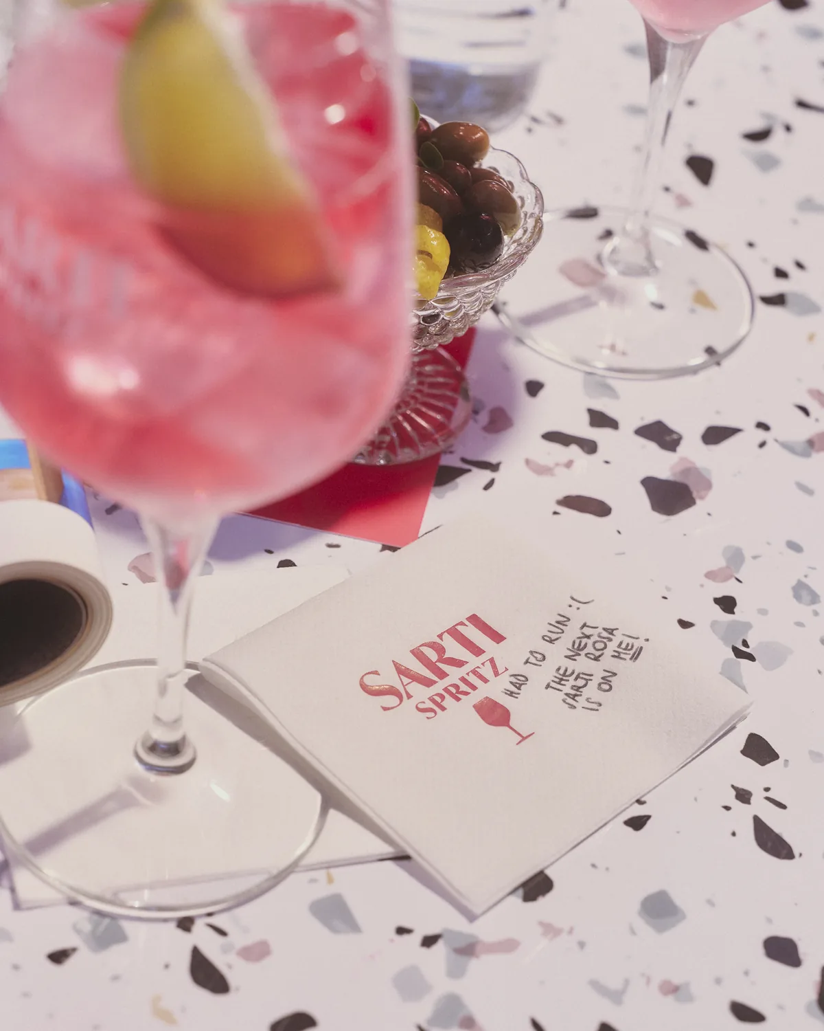 Un calice di Sarti Spritz con il lime su un tavolo alla veneziana, accanto a un tovagliolo col marchio e a una ciotola di olive